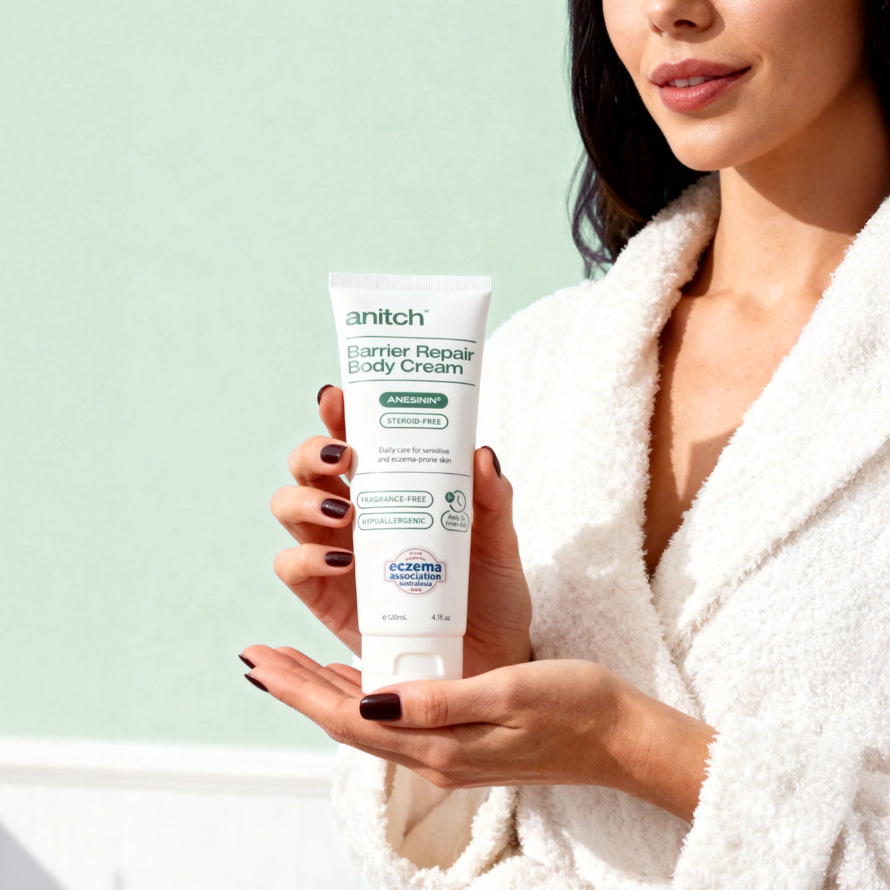 Woman holding a tube of 'anitch' Barrier Repair Body Cream against a light green background